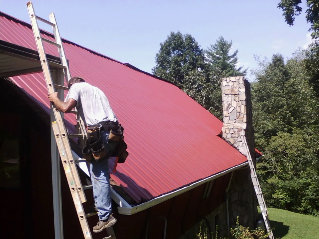 Roofer on a ladder beside a red metal roof and stone chimney — Soffit & Fascia Repair by Roofing Brothers in Granite